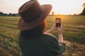 Stylish woman taking photo of sunset on phone in summer field. Atmospheric beautiful moment Royalty Free Stock Photo