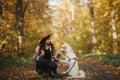 Stylish woman traveller in hat with backpack sitting with cute dog on path in sunny autumn woods. Young female hipster playing