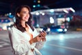 Stylish woman walking in street in dusk twilight city, using smartphone and listening music, free space