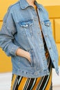 A stylish woman in a yellow skirt with diagonal lines stuck her hands in a denim jacket. Side view, right. Street style.