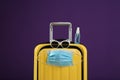 Stylish yellow suitcase with protective mask, sunglasses and antiseptic spray on purple background. Travelling during coronavirus