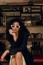 Stylish young african woman in sunglasses looking at camera sitting on bench in wine bar.