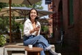 Stylish young asian woman sitting with cup of coffee in an outdoor cafe, smiling and looking happy, looking around Royalty Free Stock Photo