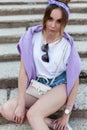 Stylish young beautiful girl in fashionable clothes with a bandana, white T-shirt, sexy jeans shorts sits on the steps and looks Royalty Free Stock Photo
