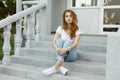 Stylish young beautiful woman in trendy summer T-shirt in fashionable jeans in trendy sneakers resting sitting on the steps Royalty Free Stock Photo