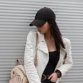 Stylish young brunette woman in a trendy white leather jacket in a trendy black baseball cap in a t-shirt Royalty Free Stock Photo