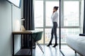 Stylish young businesswoman talking with her smartphone and looking sideways standing near the window at hotel room