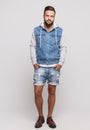 Stylish young cheerful guy with a beautiful beard in a denim jacket, a white T-shirt and denim shorts on a simple gray background Royalty Free Stock Photo