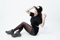 Stylish young dark-haired girl dressed in a black top, shorts, tights and cap poses sitting on the floor on the white Royalty Free Stock Photo
