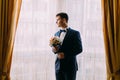 Stylish young dark haired groom holding a wedding bouquet of pale pink roses in hands standing near the window Royalty Free Stock Photo