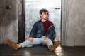 Stylish young fashion model man in bright red sunglasses and denim casual style sitting on floor and posing near metallic door and Royalty Free Stock Photo