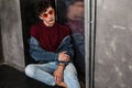 Stylish young fashion model man in bright red sunglasses and denim casual style sitting on floor and posing near metallic door and Royalty Free Stock Photo