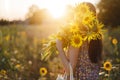 Stylish young female in floral dress walking with sunflowers in warm sunset light in summer field. Tranquil atmospheric moment in