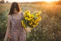 Stylish young female in floral dress walking with sunflowers in warm sunset light in summer field. Tranquil atmospheric moment in