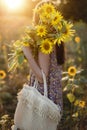 Stylish young female in floral dress walking with sunflowers in warm sunset light in summer field. Tranquil atmospheric moment in