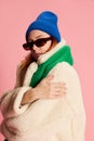 Portrait of stylish young girl in sunglasses, blue hat, green scarf and fur coat posing over pink background. Feeling Royalty Free Stock Photo