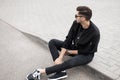 Stylish young hipster man in a black jacket in black jeans in sneakers in trendy sunglasses is sitting on the sidewalk Royalty Free Stock Photo