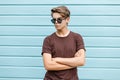 Stylish young hipster man with a fashionable hairstyle in dark sunglasses in a trendy brown t-shirt posing near a blue wooden wall Royalty Free Stock Photo