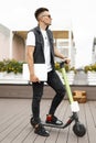 Stylish young hipster man with a modern laptop in denim black clothes in fashionable sneakers stands on a scooter on the terrace