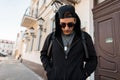 Stylish young hipster man in a trendy sweatshirt with a hood in dark sunglasses in a black cap with a stylish backpack standing Royalty Free Stock Photo