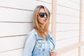 Stylish young hipster woman in black sunglasses in trendy denim blue jacket posing near a wooden vintage white building outdoors Royalty Free Stock Photo