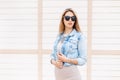 Stylish young hipster woman in black sunglasses in trendy denim blue jacket posing near a wooden vintage white building outdoors Royalty Free Stock Photo