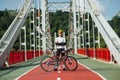 Stylish young male cyclist in full gear posing at camera with a bicycle on the bridge on a sunny day, looking at the camera Royalty Free Stock Photo