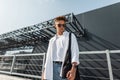 Stylish young man hipster in dark sunglasses in white fashionable clothes with hairstyle with black bag stands near a gray Royalty Free Stock Photo