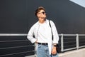 Stylish young man hipster in dark sunglasses in white fashionable clothes with trendy hairstyle with bag posing Royalty Free Stock Photo