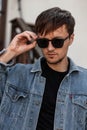 Stylish young man hipster straightens fashionable sunglasses. Portrait of a handsome guy in a trendy blue denim jacket Royalty Free Stock Photo