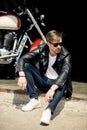 Stylish young man in leather jacket and sunglasses sitting on concrete curb near motorbike Royalty Free Stock Photo