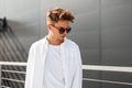Stylish young man model in vintage white shirt in trendy sunglasses with a hairstyle stands near a gray building on a sunny day. Royalty Free Stock Photo