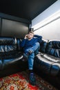 Stylish young man sitting on a black leather sofa and talking on the phone. Trendy guy in jeans and trendy sneakers makes a phone Royalty Free Stock Photo