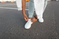 Stylish young man is standing on the pavement and tying shoelaces on leather white sneakers on a summer day. Men`s legs close-up. Royalty Free Stock Photo