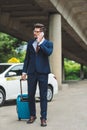 stylish young man talking by smartphone and looking away while standing with suitcase