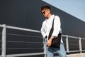Stylish young man in vintage white shirt in fashionable sunglasses trendy blue jeans with a cloth black bag posing on a sunny day Royalty Free Stock Photo