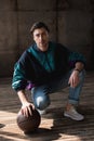 stylish young man in vintage windcheater with basketball ball sitting squats