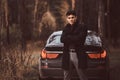 A stylish young man wearing a black coat with her arms crossed standing next to a luxury car in the autumn forest Royalty Free Stock Photo