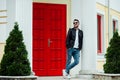 Stylish young man wearing stylish leather jacket, jeans and sunglasses posing on background of red door in sunny street. Royalty Free Stock Photo