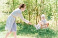 Stylish young mother swings her happy one-year-old daughter dressed in white knitted clothes on a wooden swing on a sunny day Royalty Free Stock Photo