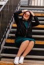 Stylish young pretty hipster woman sitting on iron house stairs, Happy beautiful girl in casual clothes green dress, black jacket Royalty Free Stock Photo