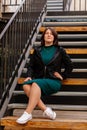 Stylish young pretty hipster woman sitting on iron house stairs, Happy beautiful girl in casual clothes green dress black jacket, Royalty Free Stock Photo