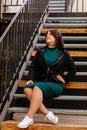 Stylish young pretty hipster woman sitting on iron house stairs, Happy beautiful girl in casual clothes green dress, black jacket Royalty Free Stock Photo