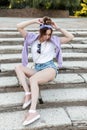 Stylish young woman with bandana in white t-shirt and ripped fashion shorts with purple sweatshirt and shoes sits on steps