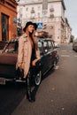 A stylish young woman in a beige coat and black hat on a city street sits on the hood of a car at sunset. Women`s street fashion.