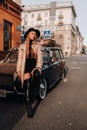 A stylish young woman in a beige coat and black hat on a city street sits on the hood of a car at sunset. Women`s street fashion.