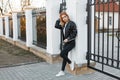 Stylish young woman in a black spring jacket in trendy jeans in a white polo shirt in sneakers posing near a vintage metal fence.