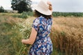 Stylish young woman in blue vintage dress and hat gathering white wildflowers in summer meadow. Beautiful girl walking with white