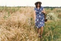Stylish young woman in blue vintage dress and hat walking with white wildflowers in straw basket at yellow wheat field. Beautiful