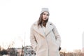 Stylish young woman fashion model in a vintage knitted hat in a fashionable faux fur coat posing at sunset outdoors. Pretty urban